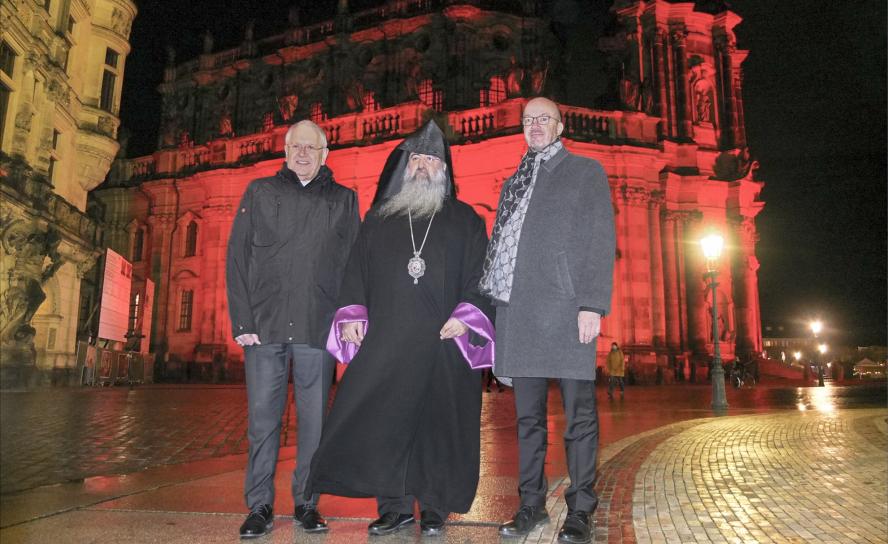 Vor der in rotes Licht getauchten katholischen Hofkirche in Dresden stehen Bischof Heinrich Timmerevers, Bischof Magar Ashkarian und Landesbischof Tobias Bilz (v. l.).Foto: Tomas Gärtner